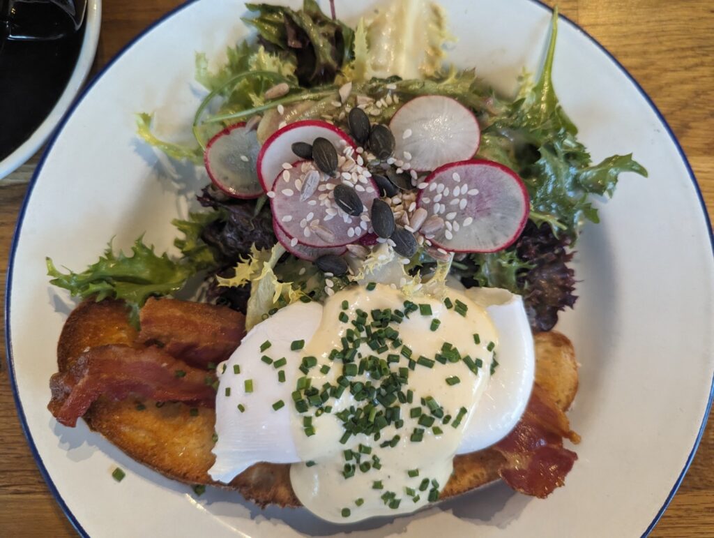Best Berlin Brunches at Oeuf restaraunt including Eggs Benadict with a poached egg, hollandaise sauce on bacon and a country bread slice served with side of salad with sliced radishes and seeds
