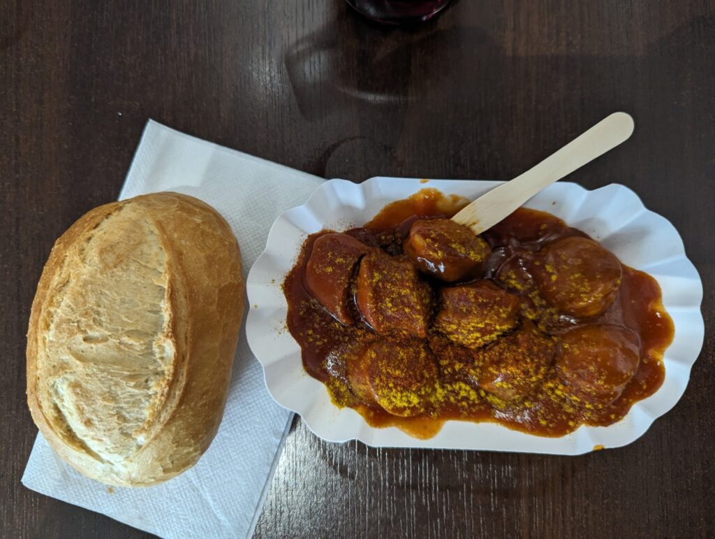 currywurst from berlin with a cut up sausage covered in a red curry ketchup and a sprinkling of yellow curry poweder on top with a roll next to the paper plate of currywurst