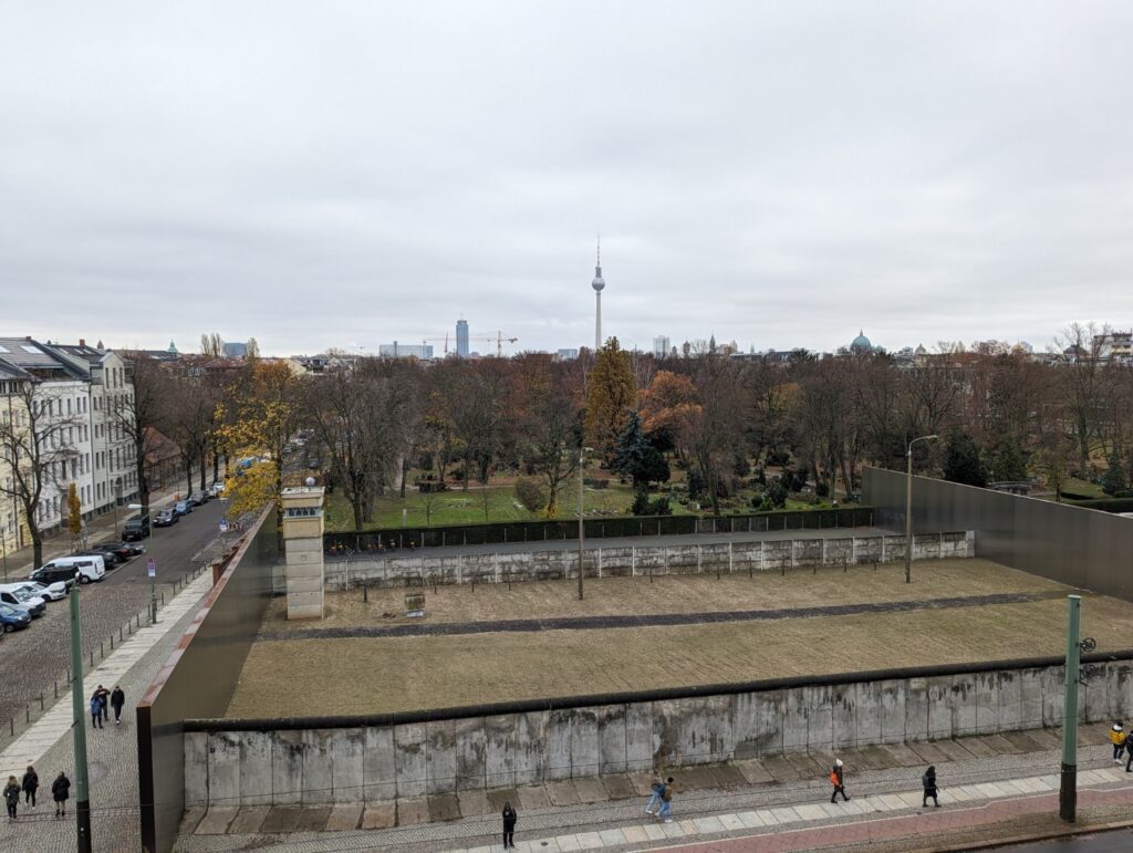 Intact section of the Berlin wall showing a grey concrete wall, the inside no man's land with a beige guard tower and then the other side of the wall. 