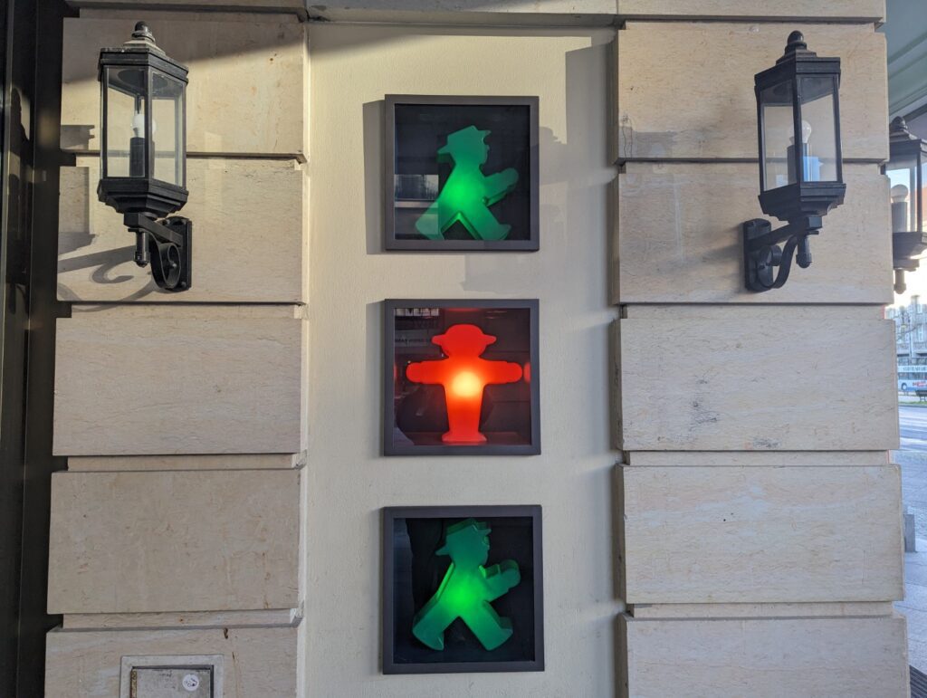 3 Berlin ampelmännchen. The top green man with a hat is the walking symbol. The Botton man has his arms outstretched and is red to symbolize no walking. The botton man is another green walking man. 